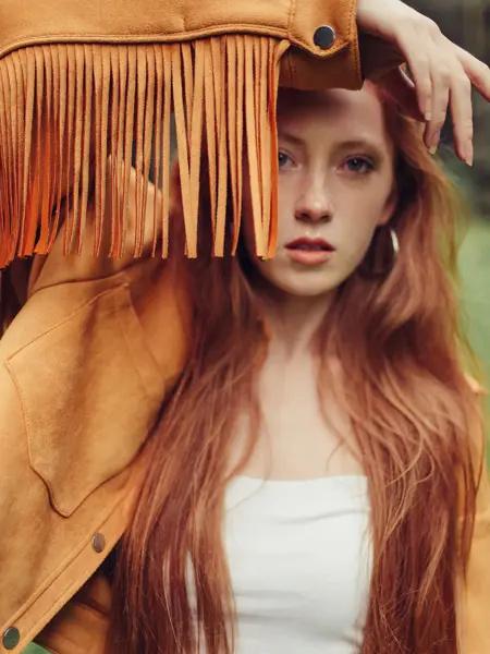 Dame med langt, rødt hår og lysebrun skinnjakke med lange frynser holder armen opp foran ansiktet, 