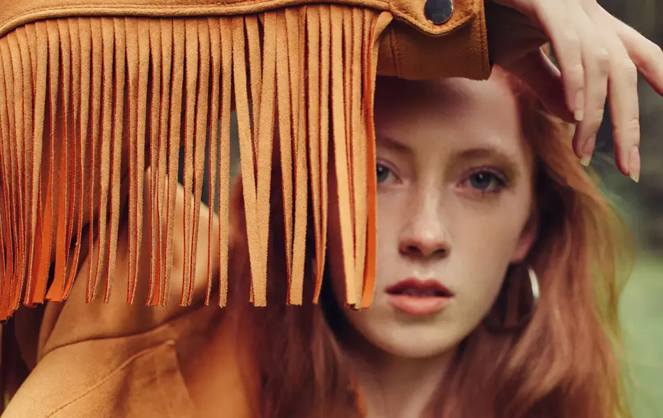 Dame med langt, rødt hår og lysebrun skinnjakke med lange frynser holder armen opp foran ansiktet, 