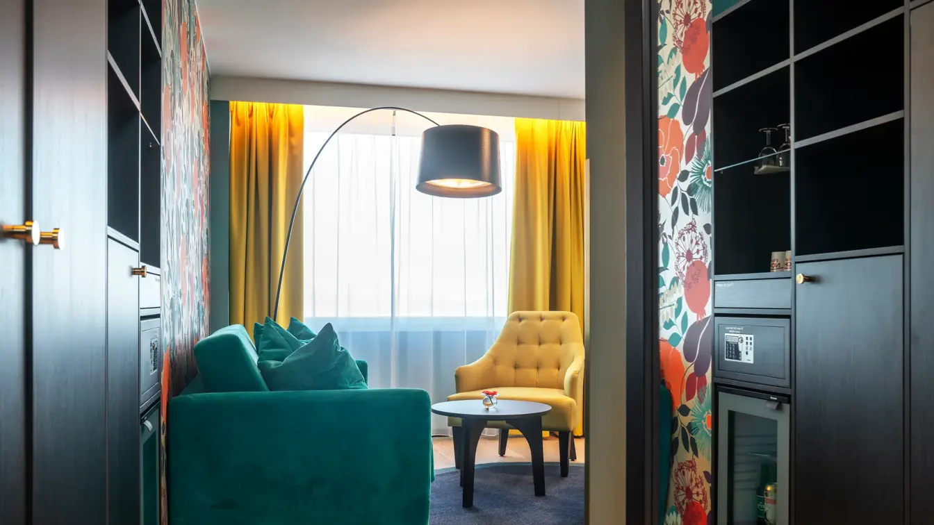 Vindu, gule gardiner, turkis sofa, skap, minibar, parkettgulv, salongbord, gul stol, gulvlampe, blomstertapetvegg
