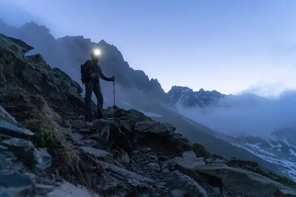 En person med hodelykt og gåstaver som går i fjellområde med dis i bakgrunnen.