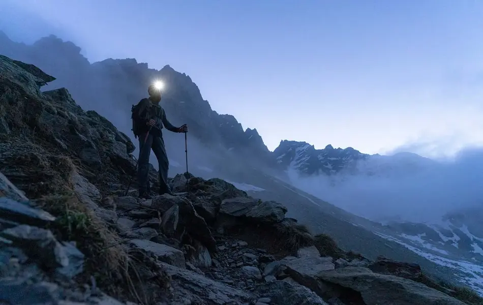 En person med hodelykt og gåstaver som går i fjellområde med dis i bakgrunnen.