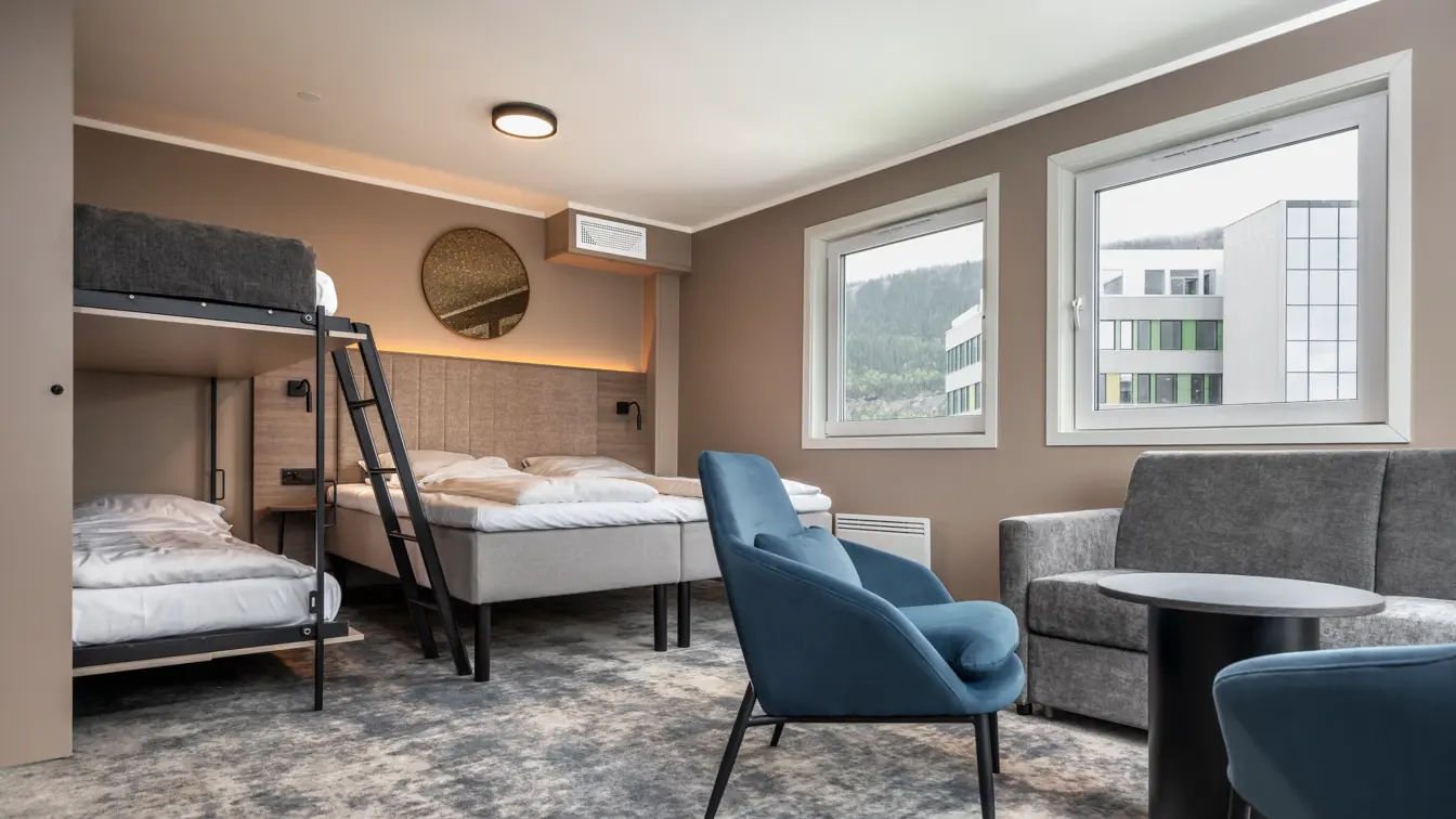 Business rom med dobbeltseng og oppredd køyeseng. Sofa, sofabord og blå lenestoler. Beige vegger og teppegulv.