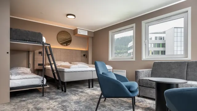 Business rom med dobbeltseng og oppredd køyeseng. Sofa, sofabord og blå lenestoler. Beige vegger og teppegulv.