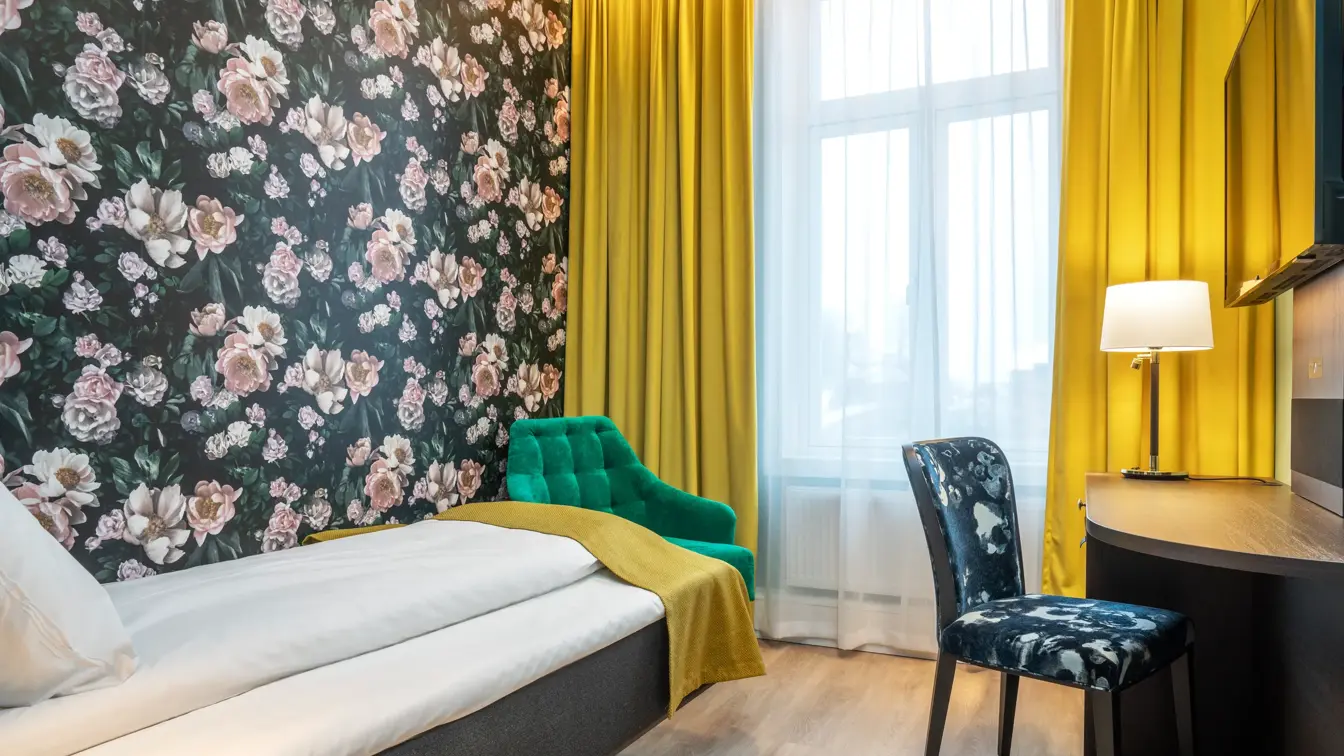 Blomster tapet med mørk bakgrunn og store lyserosa roser. Vindu med gule gardiner fra gulv til tak med en grønn lenestol foran. Enkeltseng med gult sengeteppe. Skrivebord med vegghenget TV over, mønstret stol i enkeltrom på Thon Hotel Høyers i Skien.