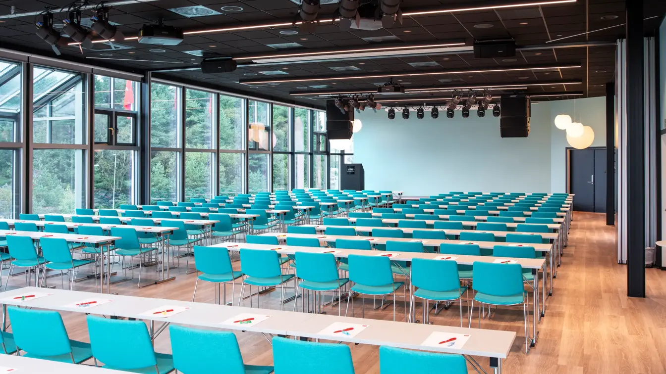 Konferansehall med skrivebord og grønne stoler. Vinduer fra gulv til tak dekker hele rommet.