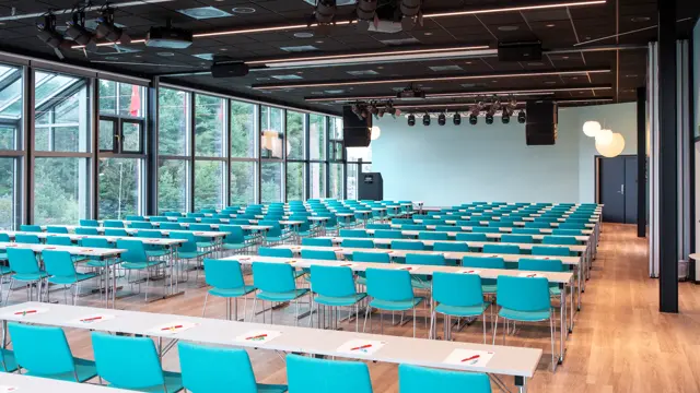 Konferansehall med skrivebord og grønne stoler. Vinduer fra gulv til tak dekker hele rommet.