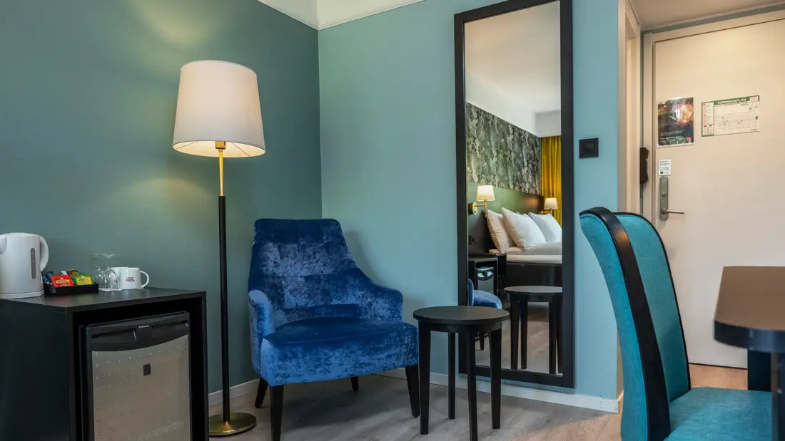 Standard rom med minibar, vannkoker, mørkeblå lenestol, lite sidebord, stålampe, skrivebord med tilhørende turkis stol og vegghengt helspeil.