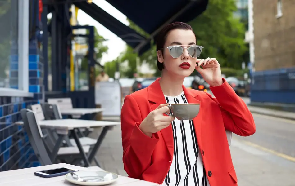 kvinne på utekafé med vertikalstripet overdel med stor, rød blazer over, solbriller, leppestift