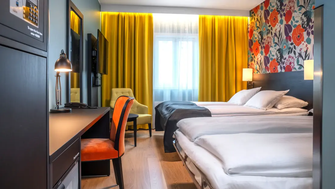 Blomstrete tapet, et stort vindu fra gulv til tak, gule gardiner, parkettgulv, fire enkle senger, svart skrivebord, en gul stol og en oransje stol. Et stort speil hengende på veggen.
