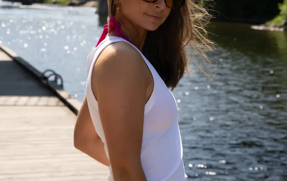 Jente på brygge med solbriller, hvit singlett, belte og blomstrete skjørt.