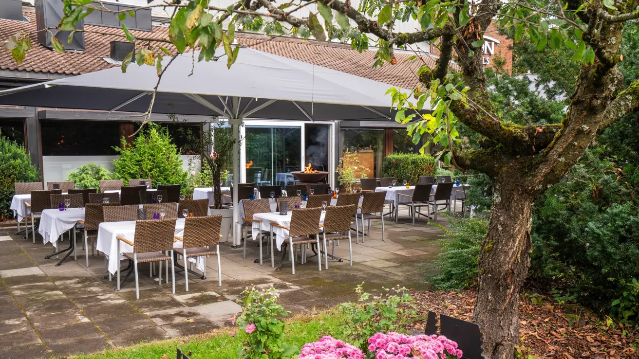 Svart blomsterkasse med blomster, stort trestående til høyre. Terasseplatting med spisebord og stoler. 