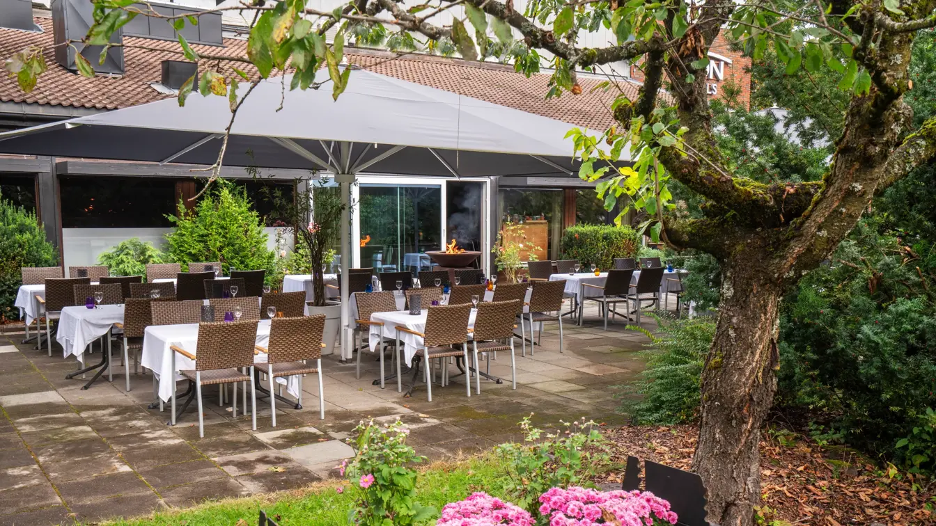 Svart blomsterkasse med blomster, stort trestående til høyre. Terasseplatting med spisebord og stoler. 