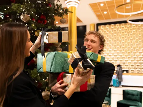 To glade personer står i lobby foran pyntet juletre med mange innpakkede julegaver i forskjellige farger.