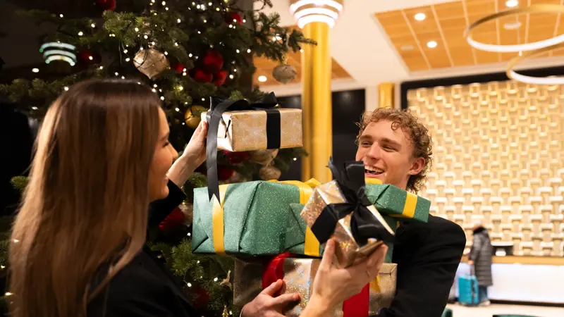 To glade personer står i lobby foran pyntet juletre med mange innpakkede julegaver i forskjellige farger.