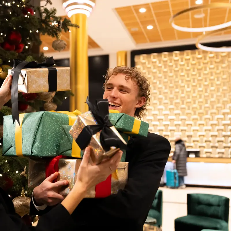 To glade personer står i lobby foran pyntet juletre med mange innpakkede julegaver i forskjellige farger.