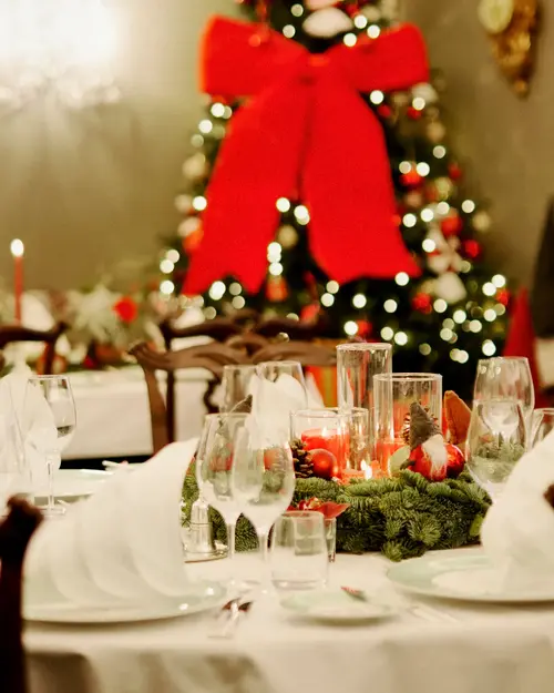 Dekket langbord med julepynt og juletre i bakgrunnen med stor rød sløyfe