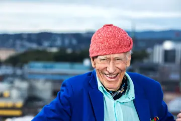 Olav Thon, med signaturluen på, smiler med Oslo-bygninger i bakgrunnen.