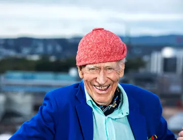 Olav Thon, med signaturluen på, smiler med Oslo-bygninger i bakgrunnen.