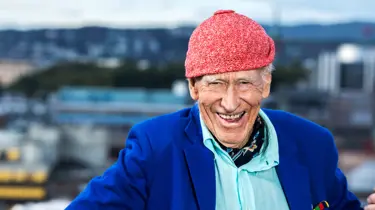 Olav Thon, med signaturluen på, smiler med Oslo-bygninger i bakgrunnen.