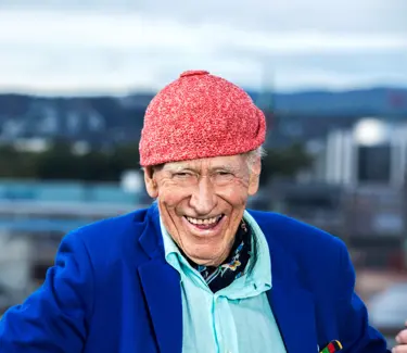 Olav Thon, med signaturluen på, smiler med Oslo-bygninger i bakgrunnen.