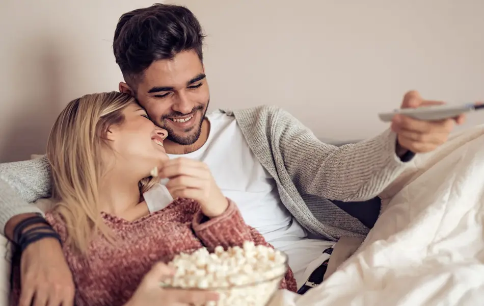 Blond jente holder skål med popkorn og lener seg på mørkhåret gutt som holder fjernkontroll