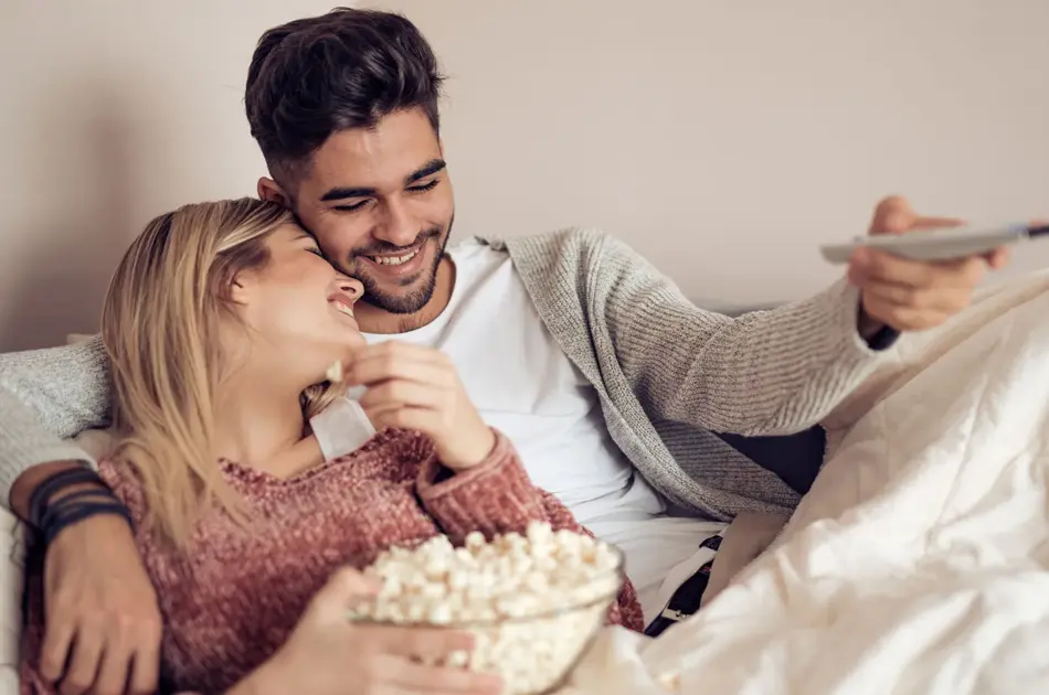 Blond jente holder skål med popkorn og lener seg på mørkhåret gutt som holder fjernkontroll