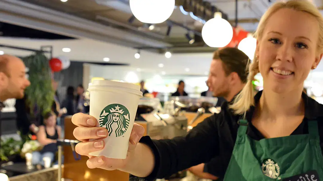 Blonde dame barista som smiler med en hvit papir kopp i hånden