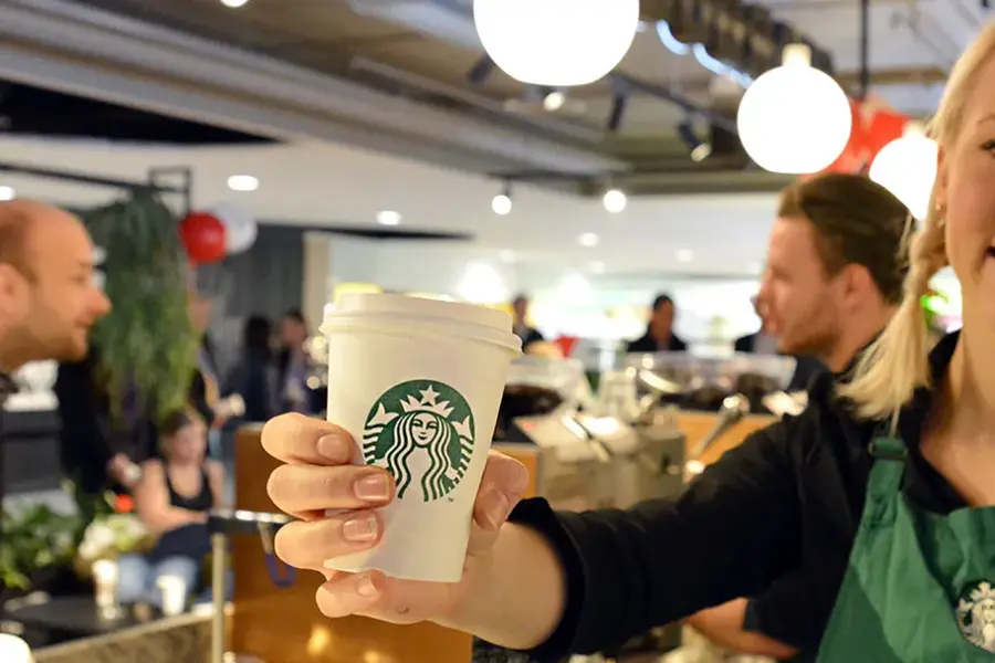 Blonde dame barista som smiler med en hvit papir kopp i hånden