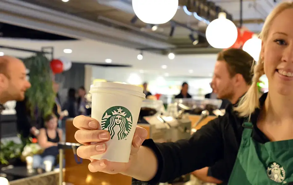 Blonde dame barista som smiler med en hvit papir kopp i hånden