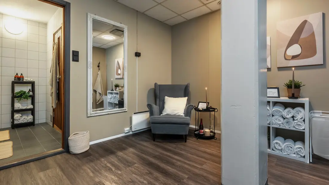 Oppholdsrom utenfor sauna med grå lenesol, håndklær, stort speil og inngang til felles dusj.