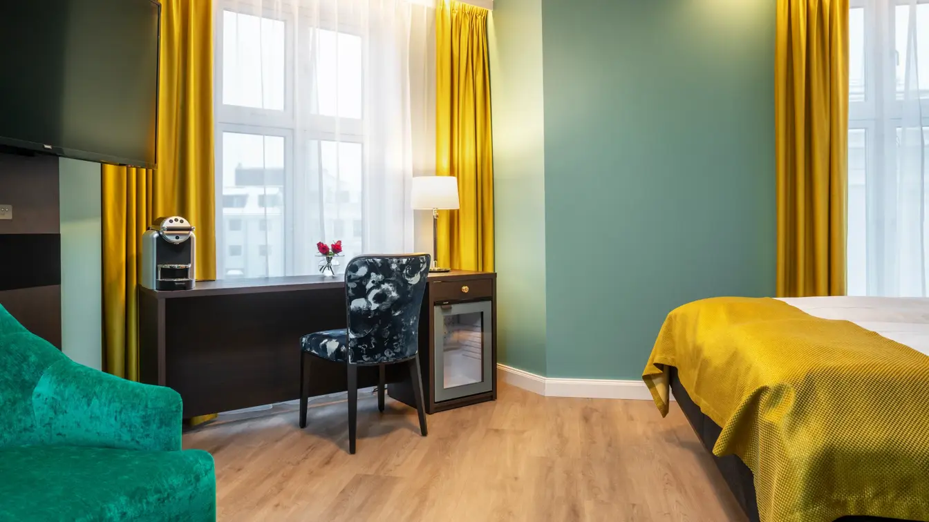 Store vinduer med gule gardiner fra tak til gulv. Mørkt skrivebord med kaffemaskin, minibar og mørk mønstret stol. Veggehengt TV og grønn lenestol. I superior rom på Thon Hotel Høyers i Skien.