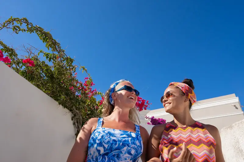 To damer med hodetørkle som matchet antrekket og solbriller står utendørs foran hvit murvegg med rosa blomster 