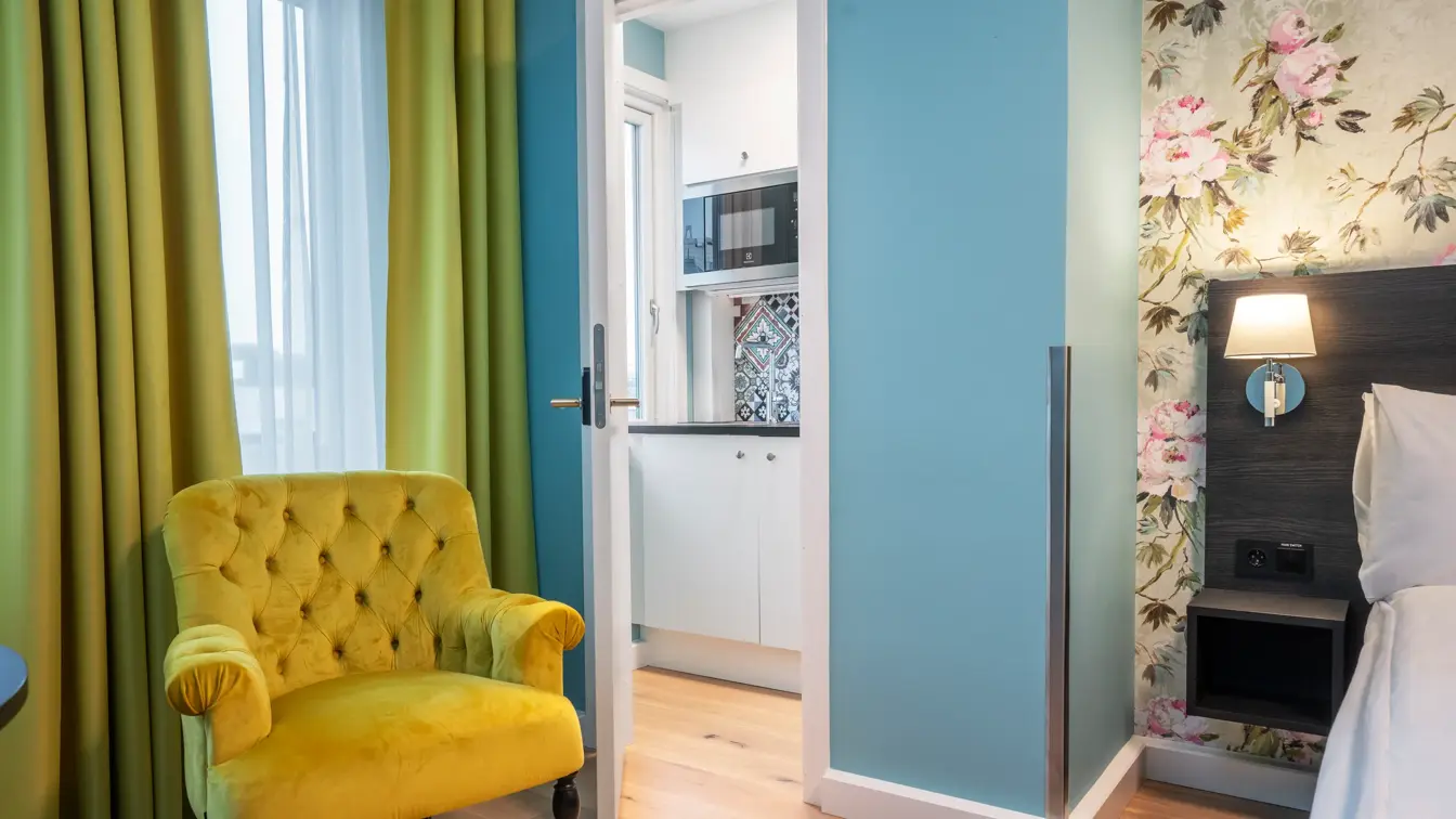 Gule gardiner, gul stol, blomstrete tapetvegg, parkettgulv, dør inn til kjøkken, dobbeltseng. 