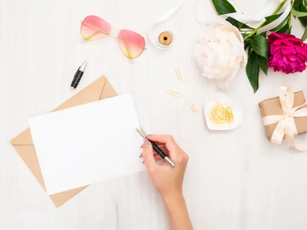 Top view woman hand writing wedding invitation card or love letter. Minimal flat lay composition with girls hand holding pen, bouquet of pion flowers, sunglasses, gift box. Writing letter concept