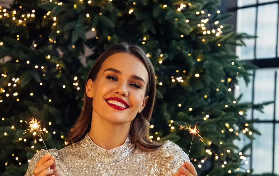 Dame med lysegrå paljettkjole sitter foran et juletre med to tente stjerneskudd og smiler til kamera