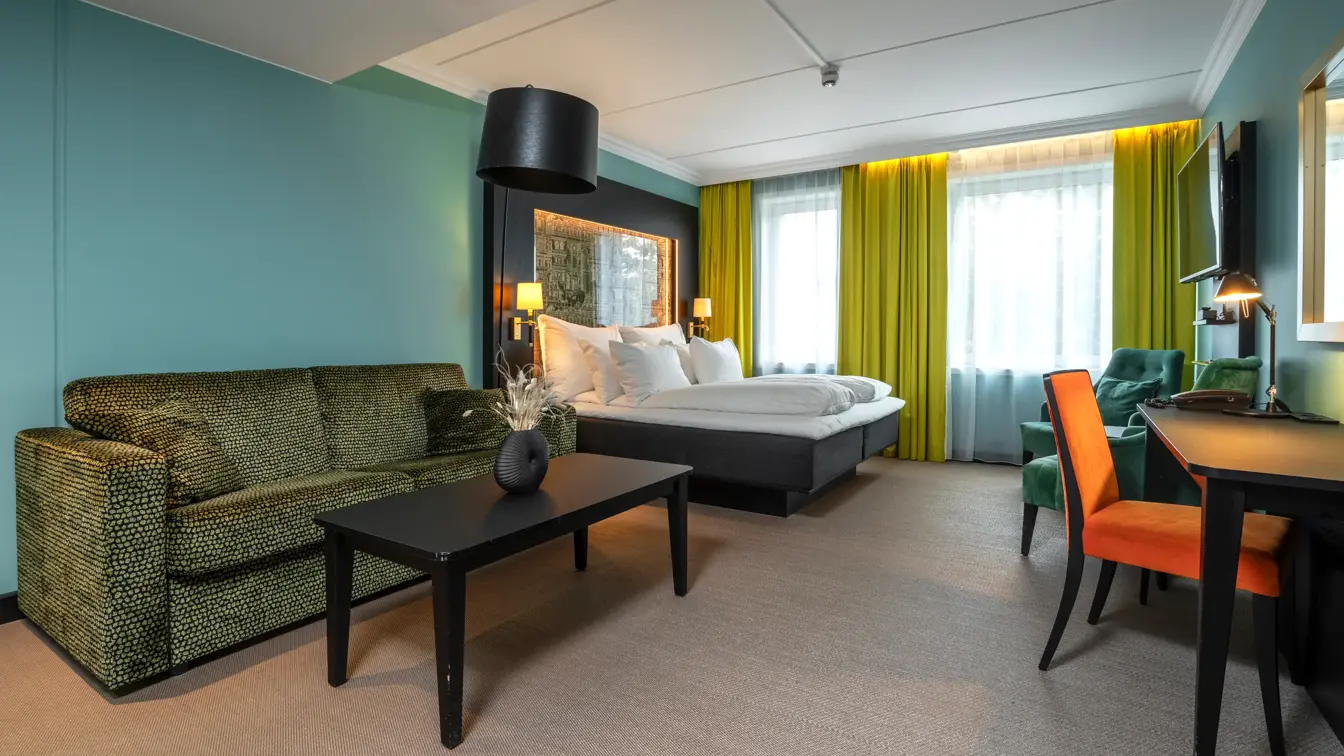 Businessrom med dobbeltseng, teppegulv, mønstrete sofa, svart salongbord, skrivepult, oransje stol og vegghengt TV.