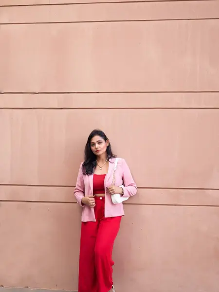 Jente med røde bukser, rød topp og rosa blazer står lent inntil en murvegg og ser mot kamera.