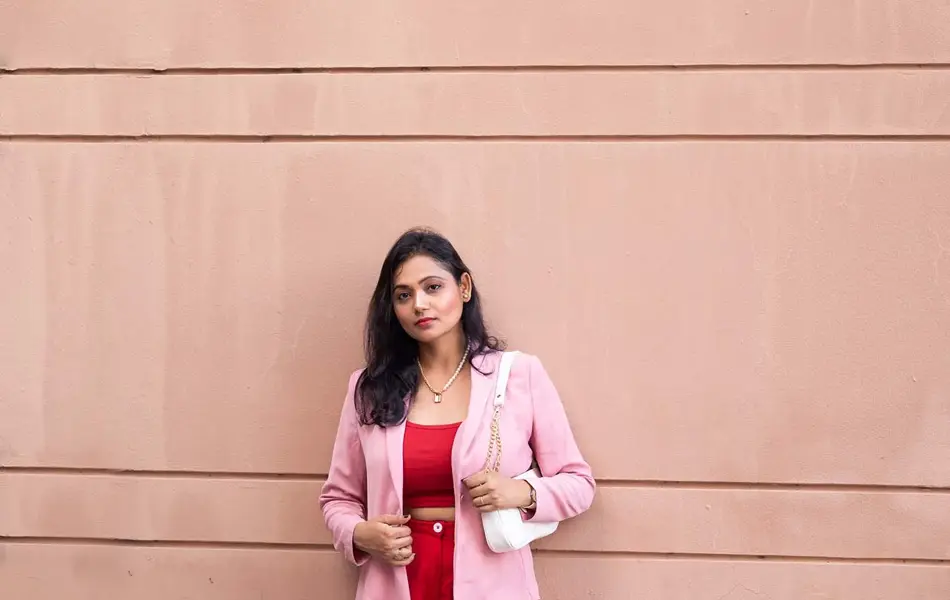 Jente med røde bukser, rød topp og rosa blazer står lent inntil en murvegg og ser mot kamera.