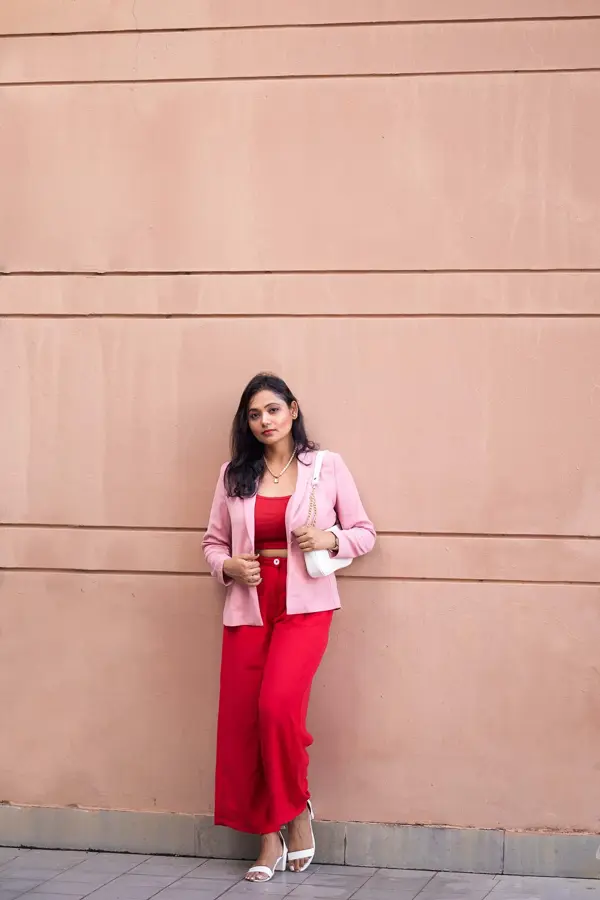 Jente med røde bukser, rød topp og rosa blazer står lent inntil en murvegg og ser mot kamera.