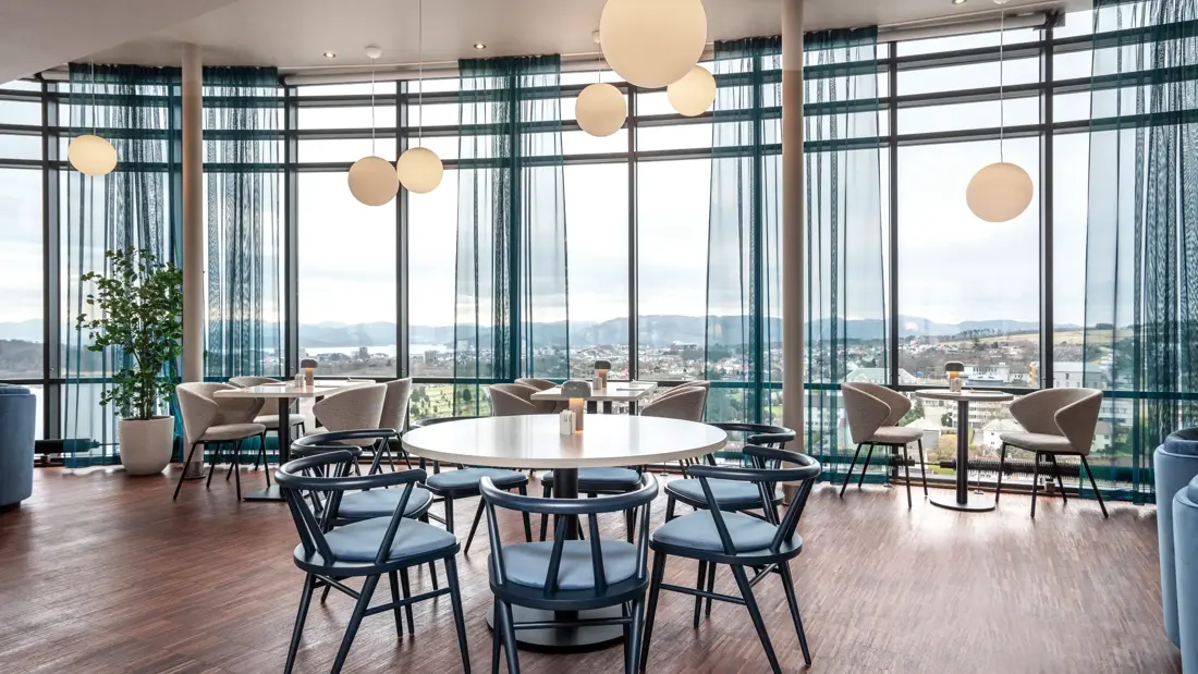 Restauranten med gulv-til-tak-vinduer på hele veggen, blå eller grå stoler rundt runde eller firkantede bord, parkettgulv.