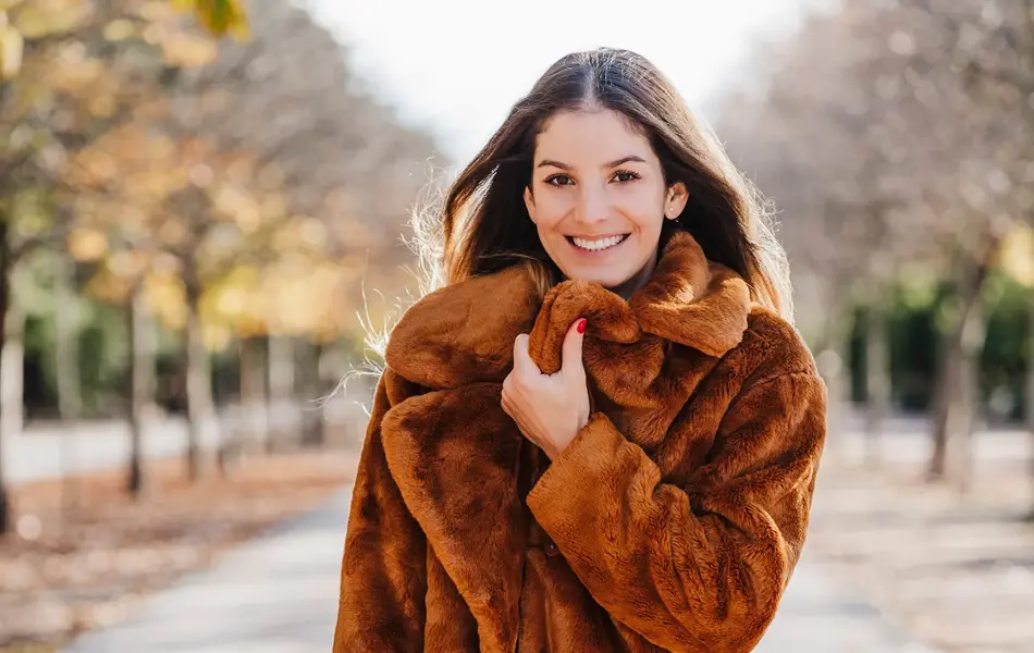Dame med lang brun fuskepelskåpe smiler til kamera på gaten med høsttrær i bakgrunnen