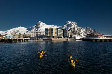 Personer padler i to kajakker i retning et havneområde med store, snødekte fjell og blå himmel i bakgrunnen.