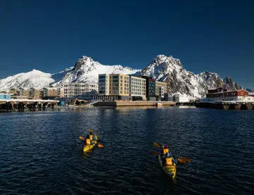 Personer padler i to kajakker i retning et havneområde med store, snødekte fjell og blå himmel i bakgrunnen.