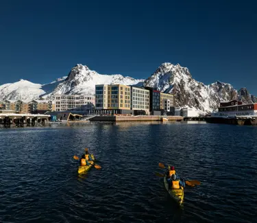 Personer padler i to kajakker i retning et havneområde med store, snødekte fjell og blå himmel i bakgrunnen.