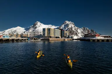 Personer padler i to kajakker i retning et havneområde med store, snødekte fjell og blå himmel i bakgrunnen.