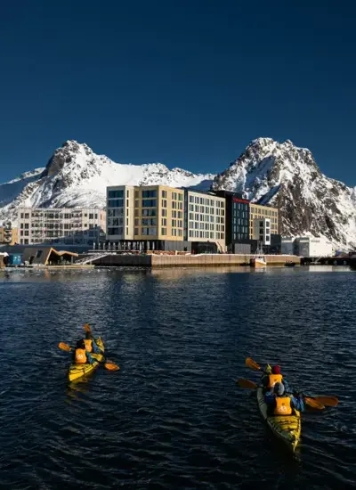 Personer padler i to kajakker i retning et havneområde med store, snødekte fjell og blå himmel i bakgrunnen.