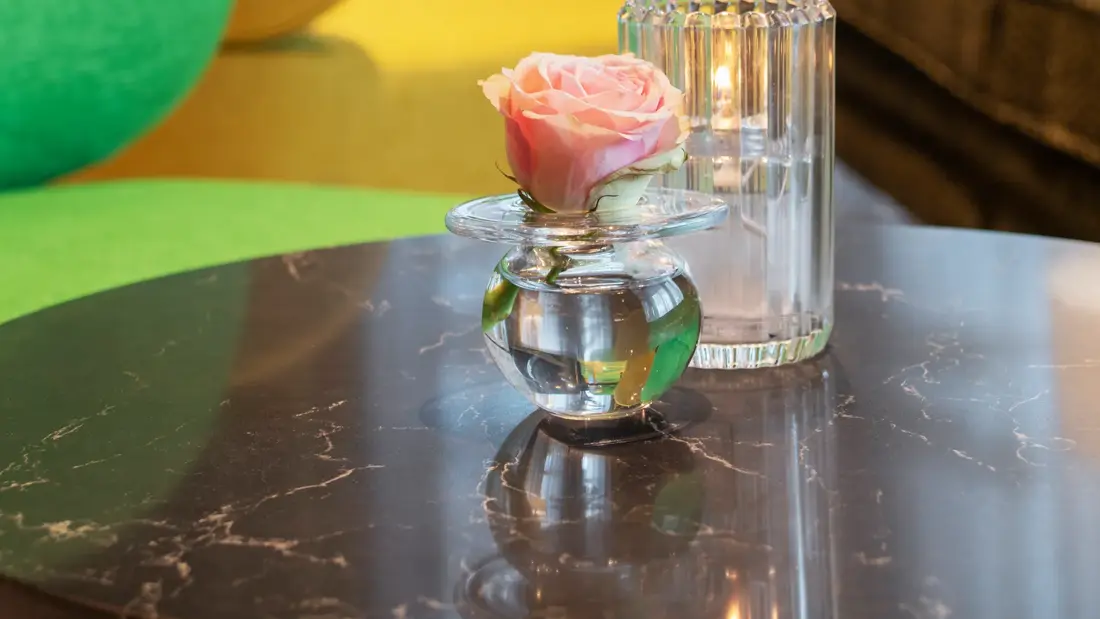 Resepsjon detalj, tre stoler i fargene gul og grønn, et lite bord med en glassvase med en rose i. Et stort vindu i bakgrunnen med en mørk sofa og en stor plante.