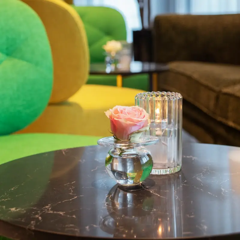 Resepsjon detalj, tre stoler i fargene gul og grønn, et lite bord med en glassvase med en rose i. Et stort vindu i bakgrunnen med en mørk sofa og en stor plante.