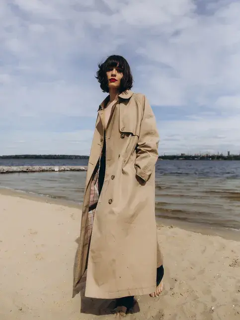 Dame med mørkt, kort hår poserer på strand med stor, beige trenchcoat 