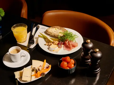 En frokosttallerken med grovt brød med smør, spekeskinke, ost, pære, valnøtter og salat. Ved siden av er et glass appelsinjuice, en kopp espresso og en tallerken med ulike oster.